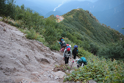 早月小屋を見下ろしながらロープの張られた上部の登山道を登っている写真