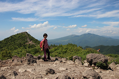 烏帽子岳の山頂と四阿山を背に稜線に立つＫちゃんの写真
