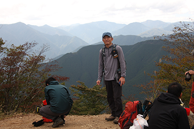 雲取山を背に山頂に立つＣさんの写真