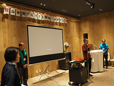 挨拶をしている理事長の写真、後には「祝山仲間アルプ１５周年！おめでとう」の横断幕がある