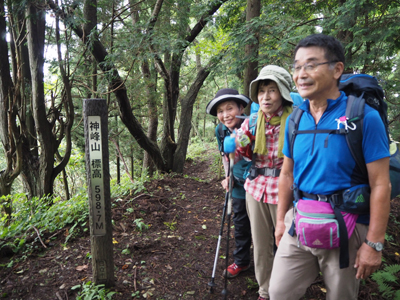 Iさんが撮影した山頂標識の横に立つメンバーの写真