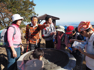 山頂の方位盤の山の方向を指さしているFさんの写真